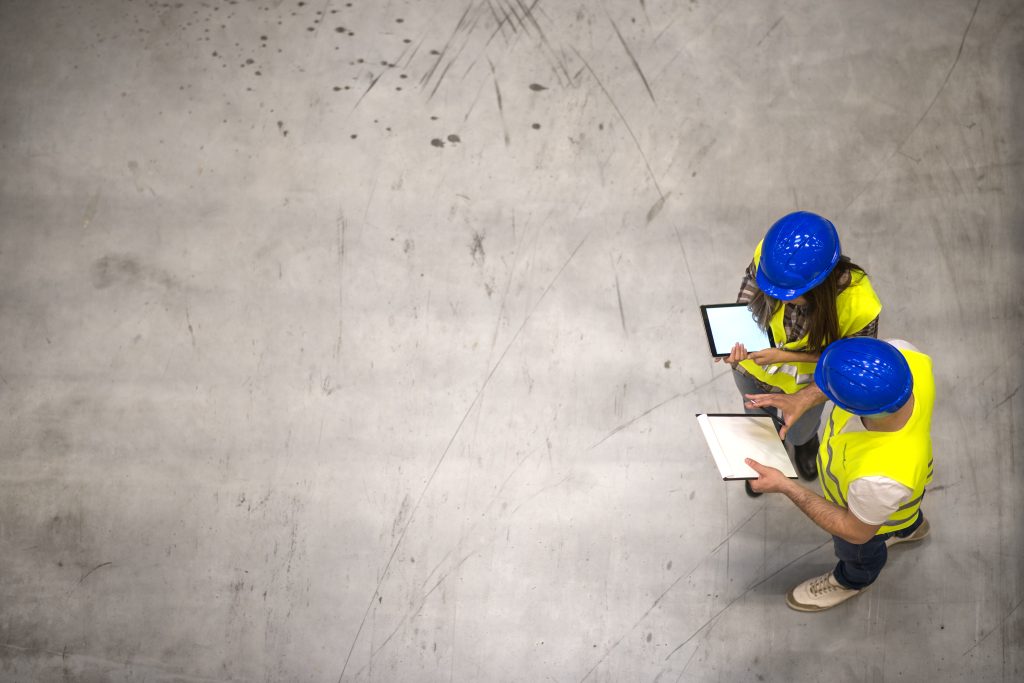 Top view of two industrial workers wearing hardhats and reflective jackets holding tablet and checklist on gray concrete background.